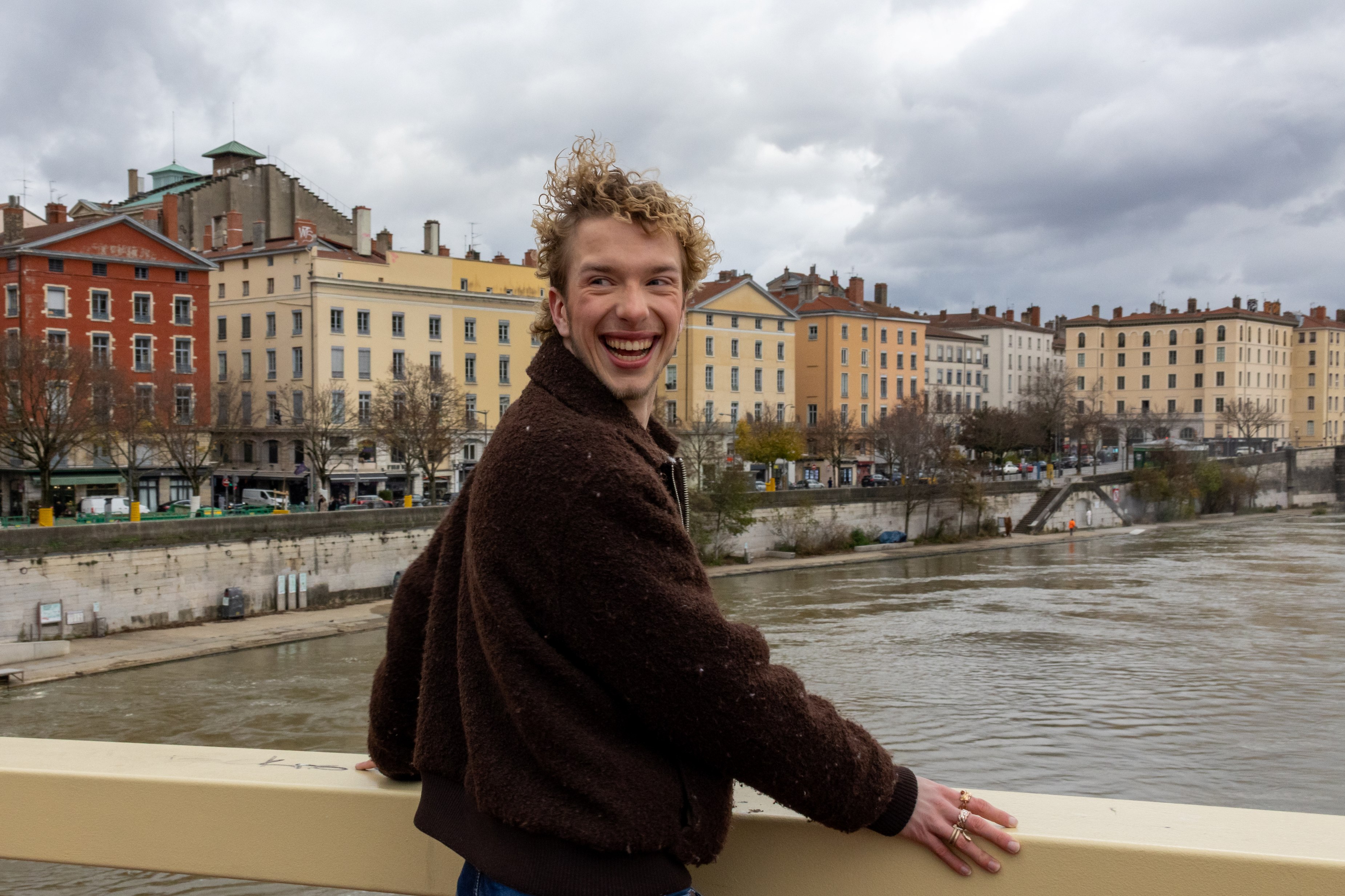 En ung mann med krøllete hår står på en bro. Han smiler. 