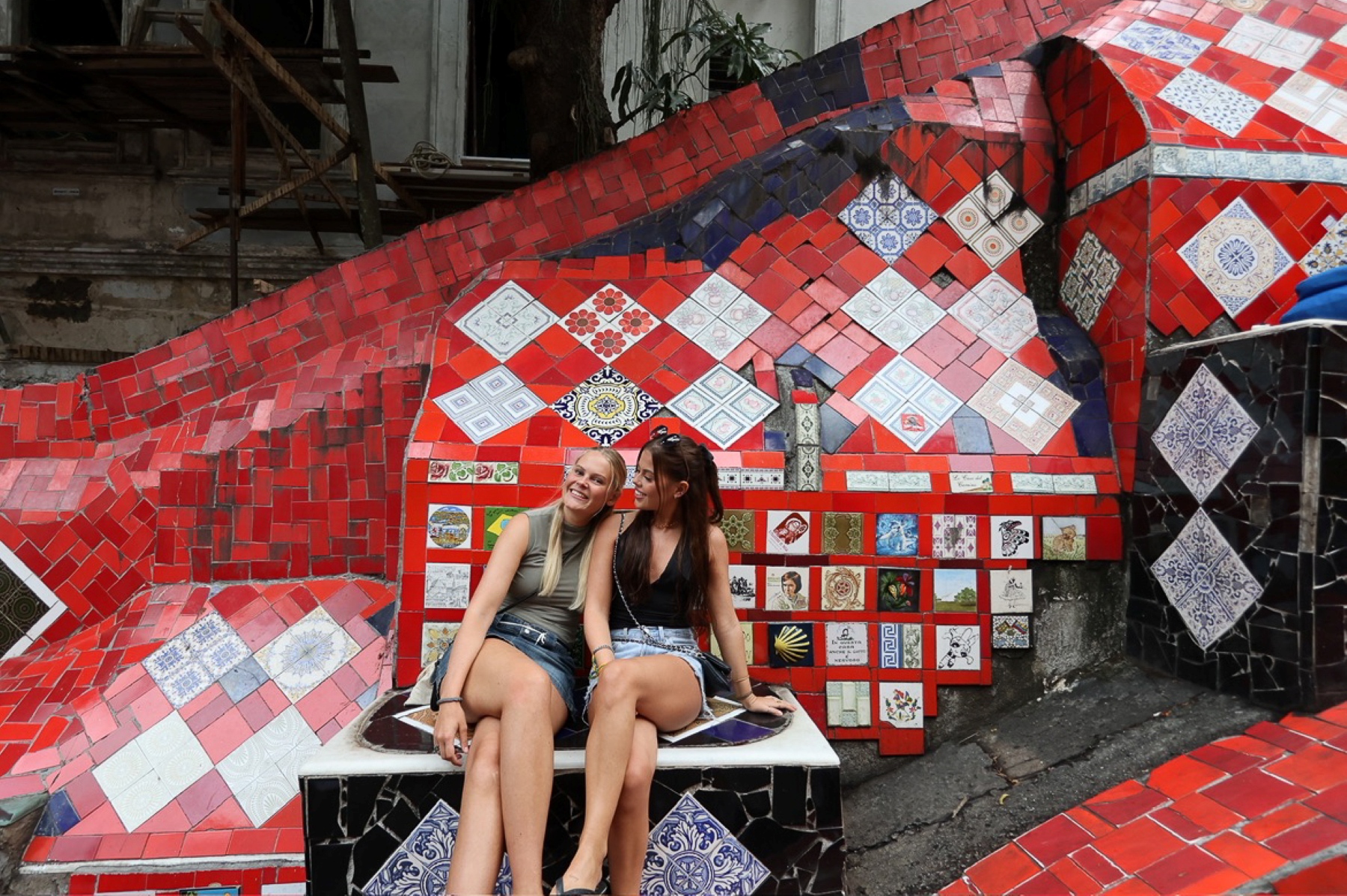 Kvinne med langt blondt hår kledd i grønnbrun topp og jeansskjørt sitter sammen med klassevenninne med svart topp og lyseblått jeansskjørt. De sitter på Escadaria Selarón, den berømte keramikktrappen i Rio de Janeiro, Brasil.