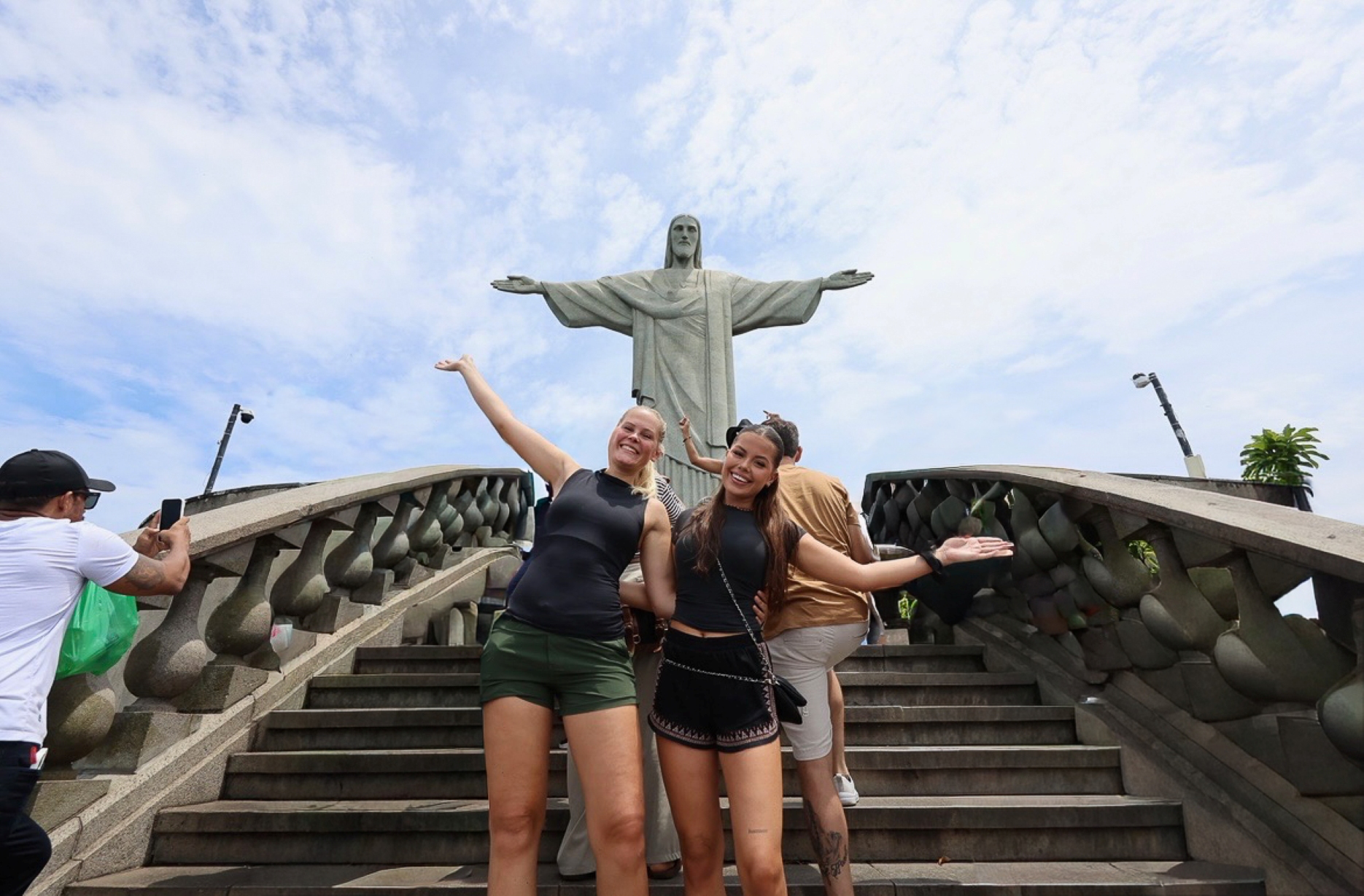 Kvinne med langt blondt hår, mørk shorts og mørk T-skjorte står sammen med venninne med brunt hår iført svart topp og svart skjørt. I bakgrunnen ser man Kristusstatuen i Rio de Janeiro.