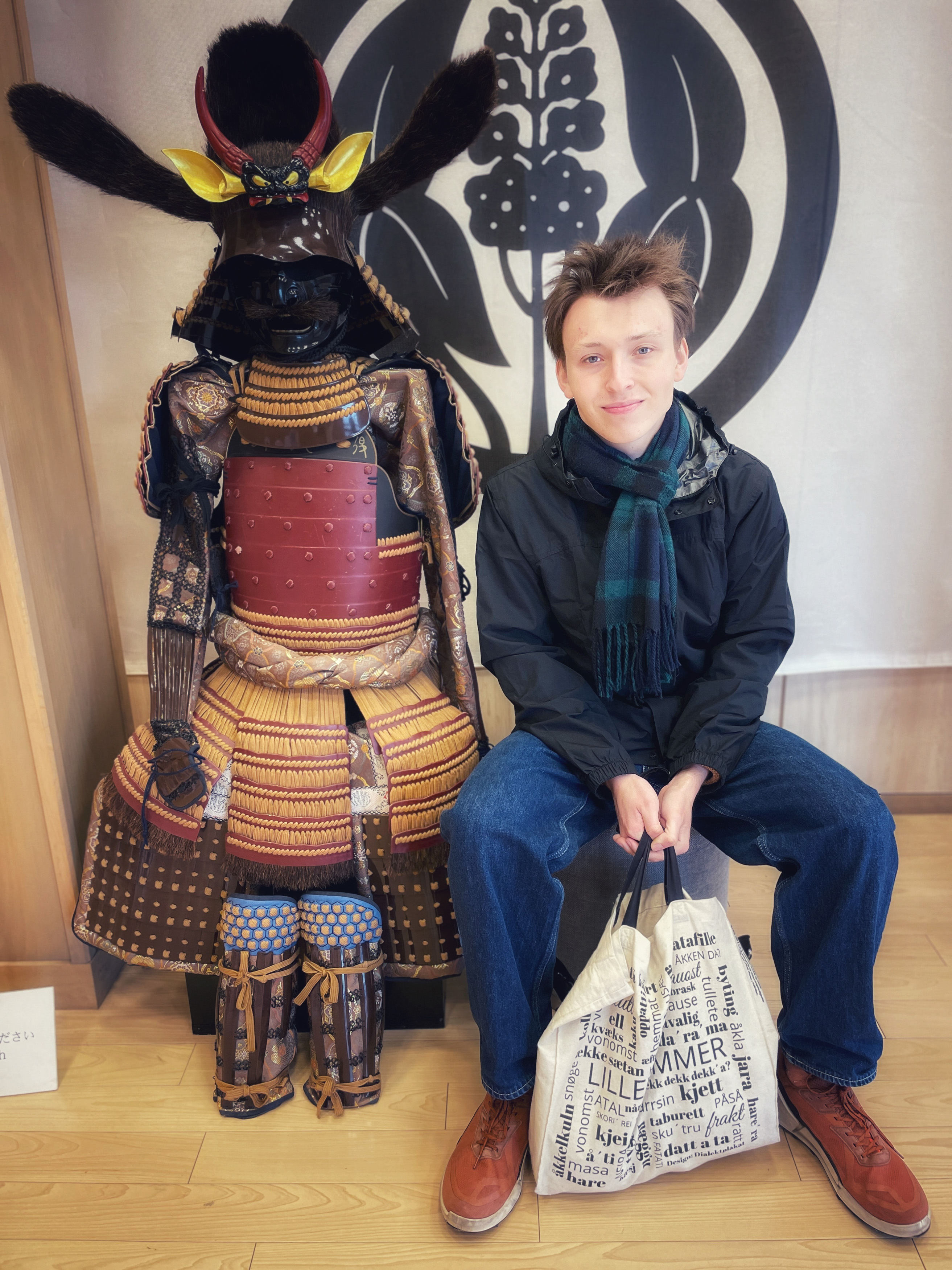 En ung mann sitter ved siden av en tradisjonell japansk samuraidraktsfigur. Han smiler. Mannen i et museum eller kulturhus.