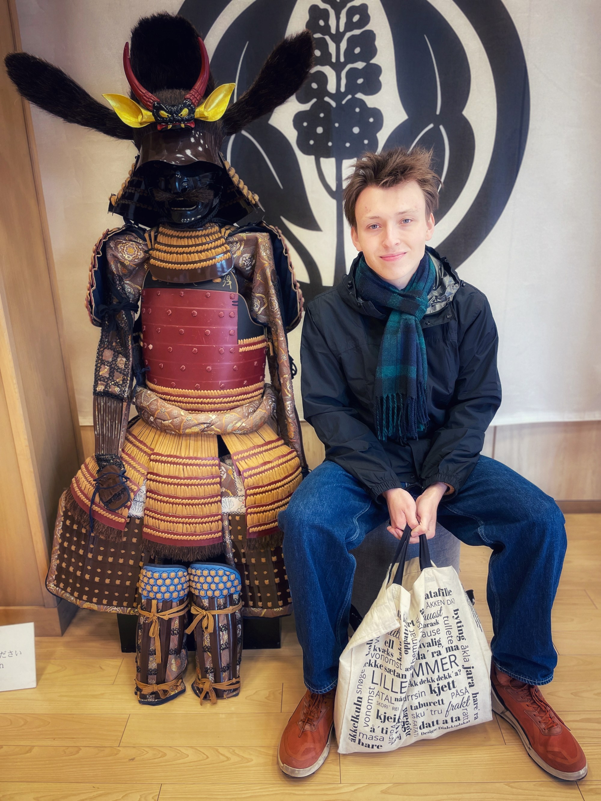 En ung mann sitter ved siden av en tradisjonell japansk samuraidraktsfigur. Han smiler. Mannen i et museum eller kulturhus.