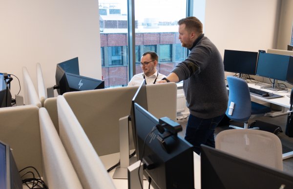 En mann i hvit skjorte og svarte briller. Han sitter foran tre pc-skjermer. Han snakker med en kollega i grå genser og mørkeblå bukser. De er i et kontorlandskap.
