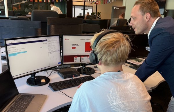 En mann står bøyd over skrivebordet til en kollega i et åpent kontorlandskap. Kollegaen sitter med ryggen til kameraet og har på seg hodetelefoner med mikrofon. På skrivebordet står flere dataskjermer som viser et salgsverktøy og nettsiden til OneCall. Rekker med små nasjonalflagg henger under taket.
