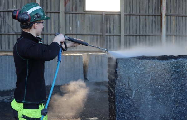 Ung mann med grønn hjelm, gule arbeidsbukser og mørk genser, høytrykksspyler en mørk steinblokk.    
