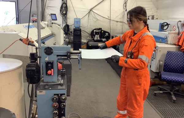 Ung kvinne i oransje kjeledress og lang mørk flette, står foran en maskin og leser en tegning.