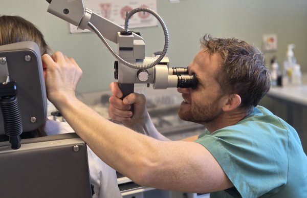 En mann ser inn i et verktøy som ligner på en kikkert, som brukes til å se inn i øret til en pasient. Pasienten sitter med ryggen til, så man ser bare at mannen holder hånda på øret til pasienten mens han ser inn i maskinen. Mannen har på seg grønn legeskjorte. 