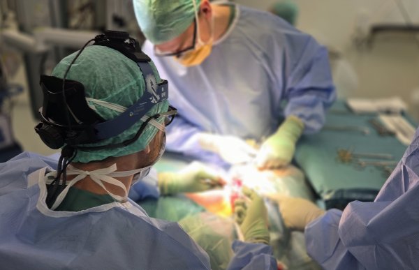 To leger gjennomfører en operasjon, de har på seg grønne hetter, briller og blå operasjonsklær og handsker. På siden er det et grønt bord med operasjonsutstyr i sølv-farge. Man ser ikke selve operasjonssåret, da det er sterkt lys der. 