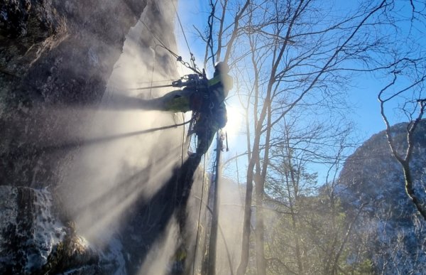 En person med verneutstyr henger i et tau og borrer i en bratt fjellside. 
