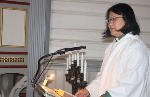 Kvinne med halvlangt svart hår og hvit kappe står på et podium, en bok ligger oppslått foran henne og hun har en mikrofon. 