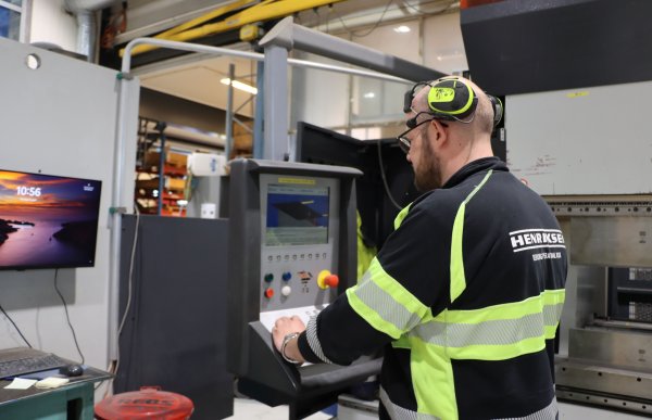 En mann kledd i svart og signalgul genser med  hørselsvern på hodet, står foran en maskin på et verksted.  