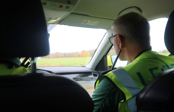 Mann med grått hår og briller sitter i forsetet på en utrykningsbil. Han har på seg en gul vest. 