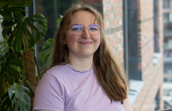 Ei dame med lyst hår og briller. Hun har på en lilla t-skjorte. Hun ser rett i kamera og smiler. Hun er på et kontor.