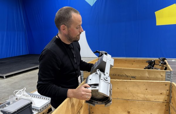 En mann i mørk genser står ved en trekasse og løfter utstyr. Bak ham er et blått stoff.