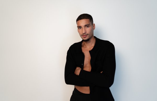 Mann med svart kort hår poserer foran kamera. Han har på seg en åpen, svart langermet skjorte og poserer foran en lys studio-bakgrunn.