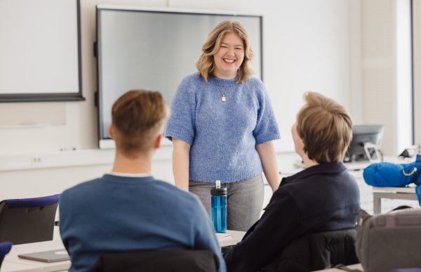 En kvinne står smilende sammen med to menn som sitter med ryggen mot kamera