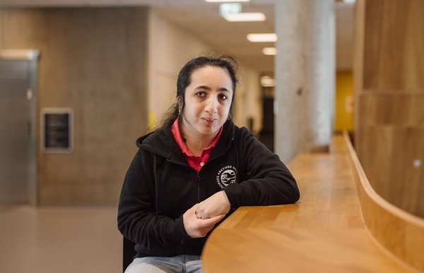 En ung kvinne med mørkt hår og svart hettejakke sitter i et fellesområde på et universitet
