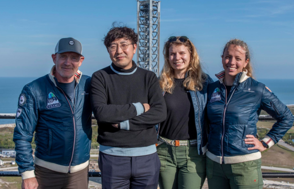 Fire personer står tett sammen og smiler foran en utsikt mot havet og en oppskytningsplattform. To av dem har på seg mørkeblå jakker med romfartsmerker, én har på seg en mørk genser, og én har solbriller på hodet. Det er sol og blå himmel, og i bakgrunnen sees kystlinje, grønt landskap og en høy metallkonstruksjon tilknyttet rakettoppskytning.