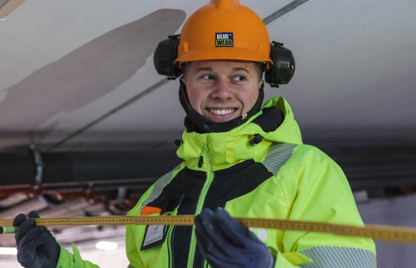 En ung mann har på seg en gulgrønn arbeidsjakke, blå hansker og oransje arbeidshjelm med hørselvern på, han holder en tommestokk og smiler mens han ser bort fra kamera