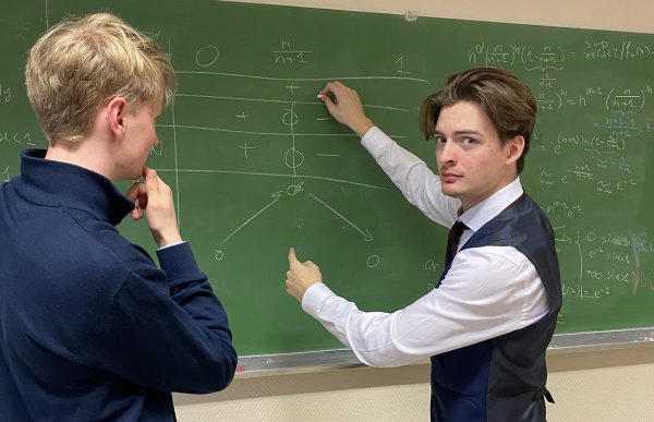 To menn står ved en tavle. Den ene peker, mens den andre følger med. Det er skrevet formler og diagram på tavlen.