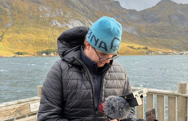 Videokunstner i arbeid i Nord-Norge Mann med blå lue og briller smiler mens han holder et kamera. Bak ham er det hav, fjell og himmel i et nordnorsk landskap.