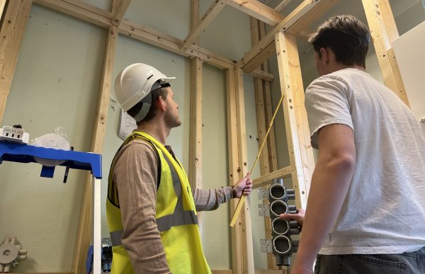 To personer står i et rom hvor reisverket i tre vises. Den ene personen har på seg gul refleksvest og hvit vernehjelm og peker en tommestokk mot den ene planken. Den andre personen har på seg hvit t-skjorte. Bilde er tatt nedenfra. De ser bort fra kamera.