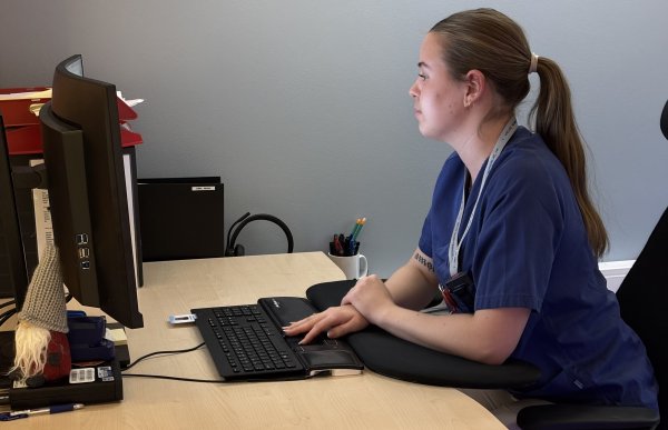 En kvinne i blå skjorte og brunt hår i hestehale sitter foran en dataskjerm 