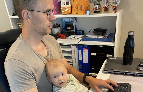 En mann sitter ved et skrivebord og jobber på datamaskin mens han holder en baby på fanget. Han har på seg en grå t-skjorte og briller og ser konsentrert på skjermen. Babyen, som ligger lent mot ham, ser rett mot kamera. På skrivebordet står en bærbar PC, tastatur, mus og en svart drikkeflaske. Bak dem står en hylle med permer, bøker, barnekunst og familiebilder, noe som gir inntrykk av et hjemmekontor.