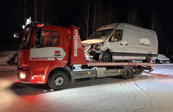 En rød redningsbil har hentet en grå varebil som har krasjet i fronten. Den grå varebilen står på et lasteplan. Det er vinter og mørkt.