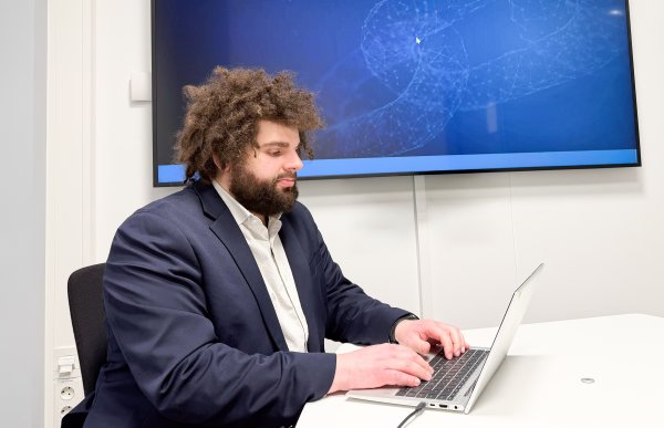 Mann med langt, brunt og krøllete hår, kledd i hvit skjorte og mørkeblå blazer, sitter på et kontor og arbeider ved en PC. I bakgrunnen vises en politi-logo på skjermen.