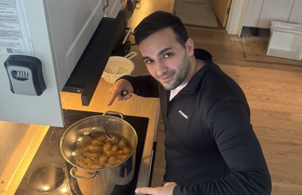 En mann i kort sort hår står ved en komfyr på et kjøkken og peker ned i en gryte med kokende mat, mens det ligger gulrøtter og epler på benken ved siden av.