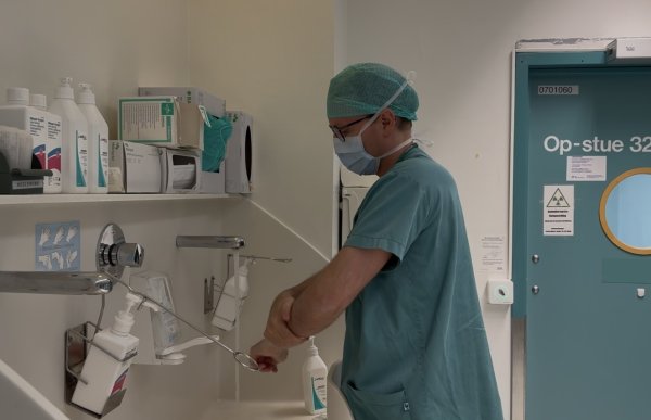 En lege i grønne sykehusklær, operasjonshette og munnbind vasker hendene ved en vask utenfor en operasjonsstue. Ved siden av vasken står det flasker med desinfeksjonsmiddel og annet hygienisk utstyr. På døren bak ham står det «Op-stue 32».