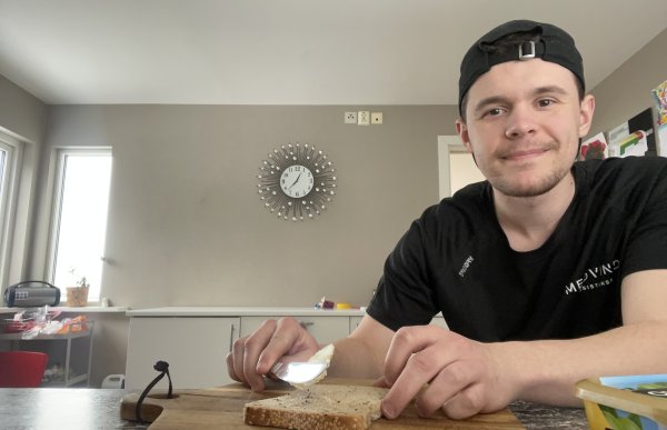 En ung mann med svart t-skjorte og svart caps sitter ved en kjøkkenbenk og smører pålegg på en brødskive