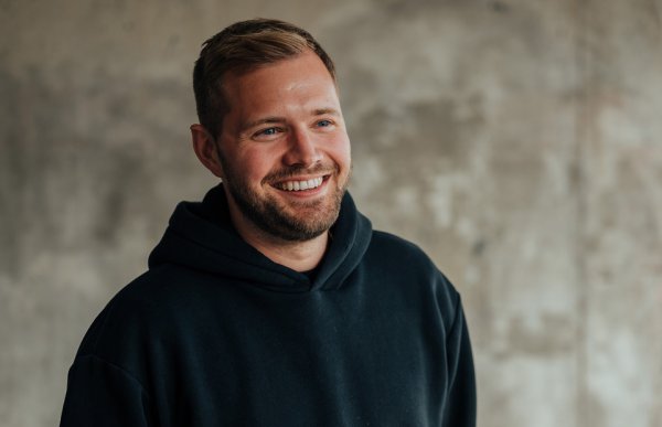 En mann med blondt hår og skjegg og svart hettejakke står foran en grå vegg