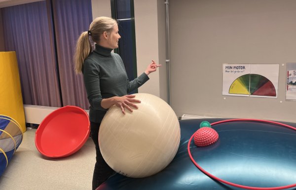 En kvinne i grå genser og med hestehale står foran en plakat og holder på en stor hvit ball