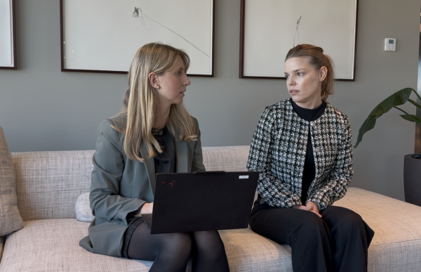 To kvinner sitter ved siden av hverandre på en lys sofa i et kontor. Kvinnen til venstre har en bærbar PC på fanget og er kledd i mørke, svarte klær med en grå dressjakke over. Hun vender seg mot kvinnen ved siden av, som har håret satt opp og har på seg en mønstret jakke over mørk topp. De ser på hverandre og ser ut til å være i samtale. Bak dem henger enkle kunstverk på veggen, og en grønn plante står i rommet.