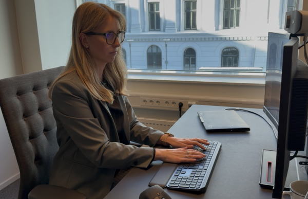 En kvinne sitter ved et skrivebord foran en dataskjerm og skriver på et tastatur. Hun har på seg briller, en svart topp og en grå dressjakke, og ser konsentrert på skjermen. På skrivebordet ligger en lukket bærbar PC, og ved siden av står en kaffekopp. Gjennom vinduet bak henne er en bygning med klassisk fasade synlig.