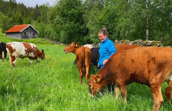 Dame med brunt hår og blå genser står på et jorde med kyr rundt seg. En grå trebygning i bakgrunnen sammen med bjørkeskog. 