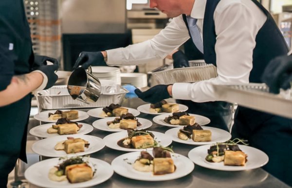 Et kjøkkenmiljø under matservering. Flere tallerkener med forseggjort mat er plassert på et langt bord. En person i kokkeuniform og en servitør i hvit skjorte og vest samarbeider om å legge saus på rettene. Stemningen er profesjonell og travel.