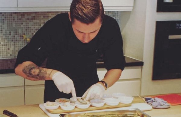 En kokk står på et profesjonelt kjøkken iført svart kokkeuniform, grå forkle og svart caps. Han arbeider konsentrert ved en benk omgitt av stålfat, boller og kjøkkenutstyr. På armen har han synlige tatoveringer, og han bruker blå hansker mens han tilbereder råvarer.