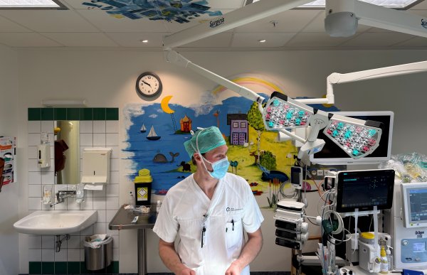 En mann i hvitt operasjonstøy, grønn operasjonshette og munnbind står inne på en operasjonsstue. Rundt ham står medisinsk utstyr, skjermer og operasjonslamper. Veggene er dekorert med fargerike tegninger av blant annet hav, regnbue og hus for å gjøre rommet mer barnevennlig.