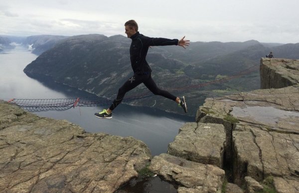 En person hopper med store steg mellom to fjellkanter på Preikestolen, med fjord og fjellandskap langt under og rundt.