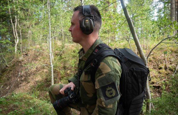 En person i militæruniform står i skogen med et kamera og hørselvern. Personen har en ryggsekk og ser utover terrenget mellom trær og lyng.