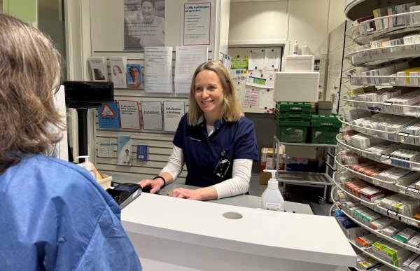 En kvinne i arbeidsuniform sitter bak apotekdisken og snakker med en kunde. Hun lener seg lett fremover og smiler. På disken ligger betalingsutstyr og hånddesinfeksjon. I bakgrunnen sees hyller med legemidler og informasjonsmateriell.