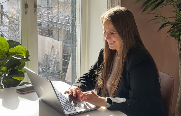 En dame sitter ved et bord og skriver på en laptop. Hun sitter ved vinduet og det er solskinn ute.