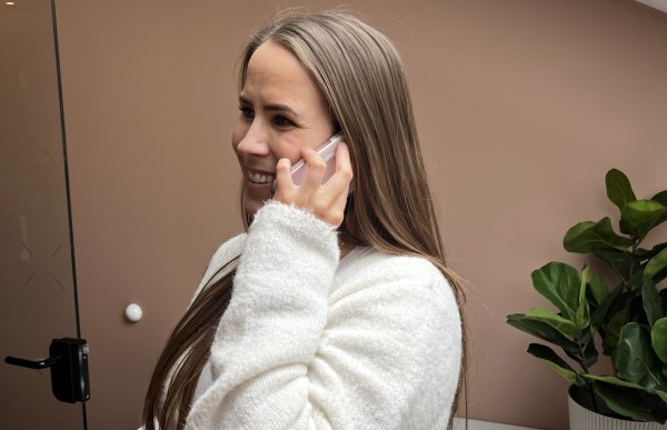 En dame holder telefonen inn til øret og smiler. I bakgrunnen er en beige vegg og en plante til høyre i bildet. Hun har på seg en hvit genser og har langt mørkeblondt hår.