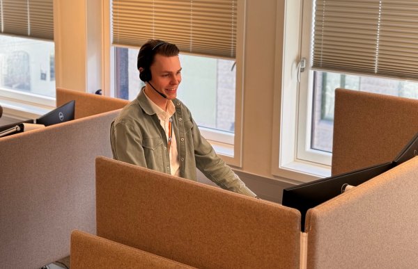 En mann i lysegrønn jakke. Han har headset på hodet. Mannen ser på en pc-skjerm. Han smiler. Han er i et kontorlandskap.