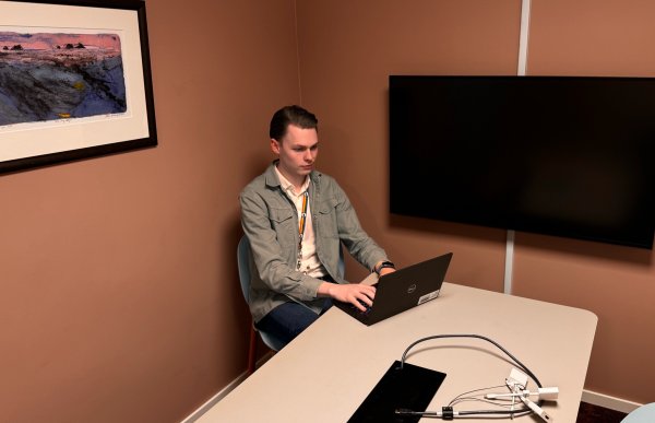 En mann sitter ved et bord og skriver på en pc. En TV henger på veggen og et bilde i en bilderamme.