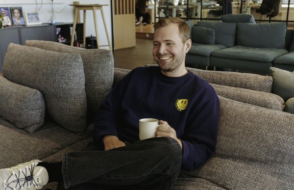 En mann i svart genser, svart bukse og hvite joggesko sitter i en grå sofa med en kaffekopp