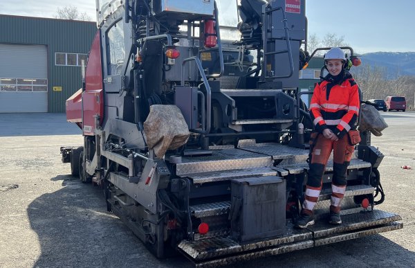 En dame står ved bakpå stor asfalteringsmaskin. Hun har på seg oransje arbeidsklær med refleks og vernehjelm. Maskinen står på en åpen plass med et grønt bygg i bakgrunnen. 