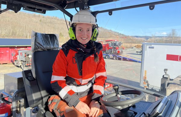 En jente sitter med vernehjelm og hørselsvern i en maskin. Hun har på seg oransje arbeidsjakke. I bakgrunnen sees andre maskiner og et skogholdt. Det er blå himmel i bakgrunnen. 
